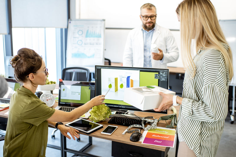 Two people working at a desk with computer monitors displaying graphs, with another person in the background.
