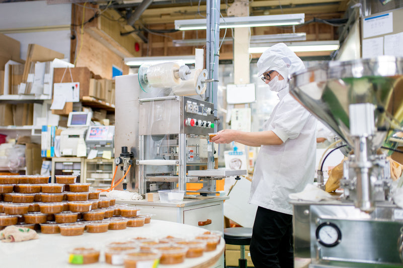 Person working in a factory setting with packaging machinery and products on shelves.