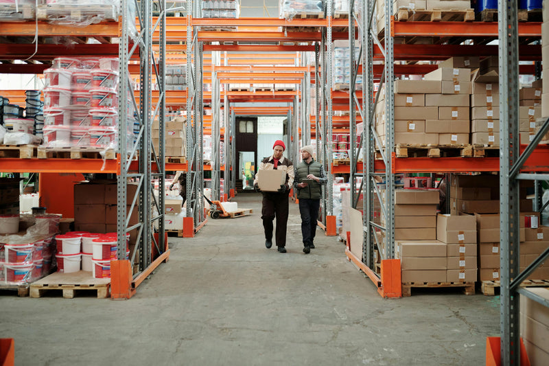 Two workers walking through a warehouse with shelves and boxes.
