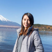 Woman in a gray coat standing in front of Mount Fuji and a lake.