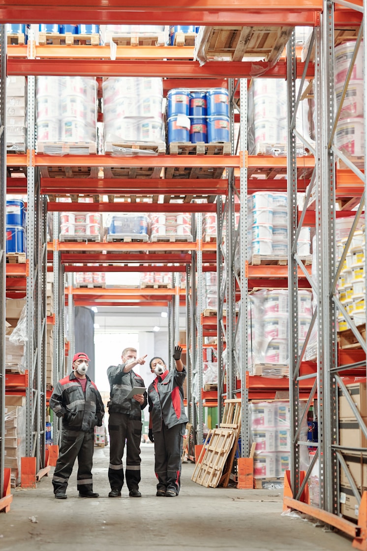 Three workers in a warehouse with high shelves and storage racks.