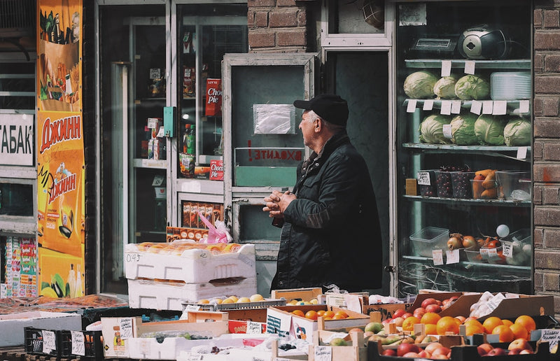 Man standing behind a fruit and vegetable stand with shelves displaying more produce.