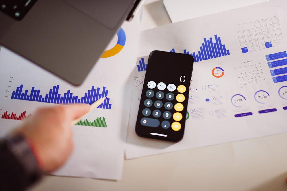 Hand using a smartphone calculator on financial documents with charts and graphs.