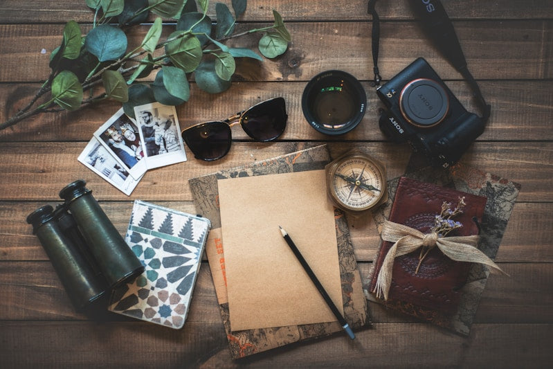 Scattered items on a wooden surface including binoculars, camera, compass, and notebook.