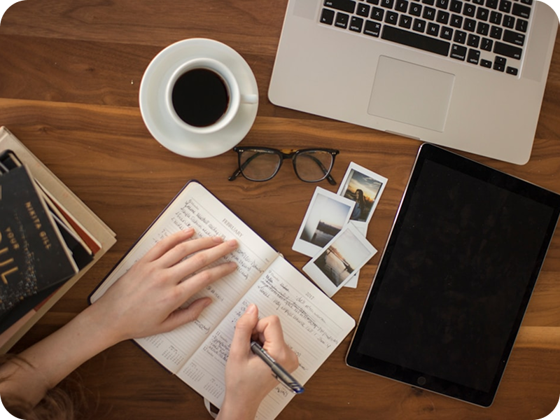 Person writing in a notebook on a wooden desk with a laptop, tablet, coffee, and photos.