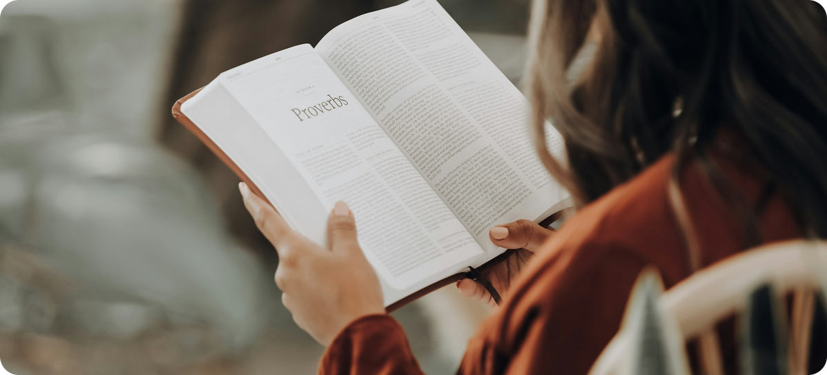 Person holding and reading an open book titled 'Proverbs' in a blurred indoor setting.