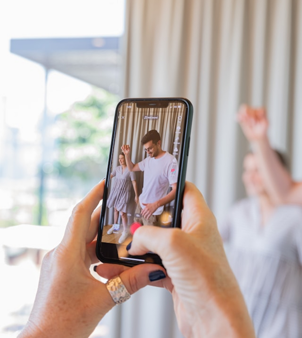 Person holding a smartphone recording a video of two people dancing in a room with large windows.